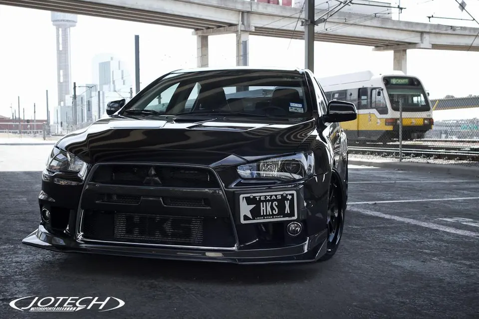 Black Mitsubishi Lancer Evolution X with HKS intercooler and front splitter in an urban setting with a train and Dallas skyline.