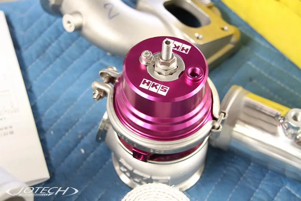 A close-up of a purple HKS blow-off valve on a blue mat, with other silver car parts in the background.