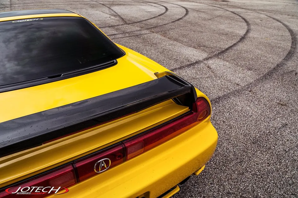 Yellow Acura NSX rear view with a carbon fiber wing on an asphalt surface with tire marks.