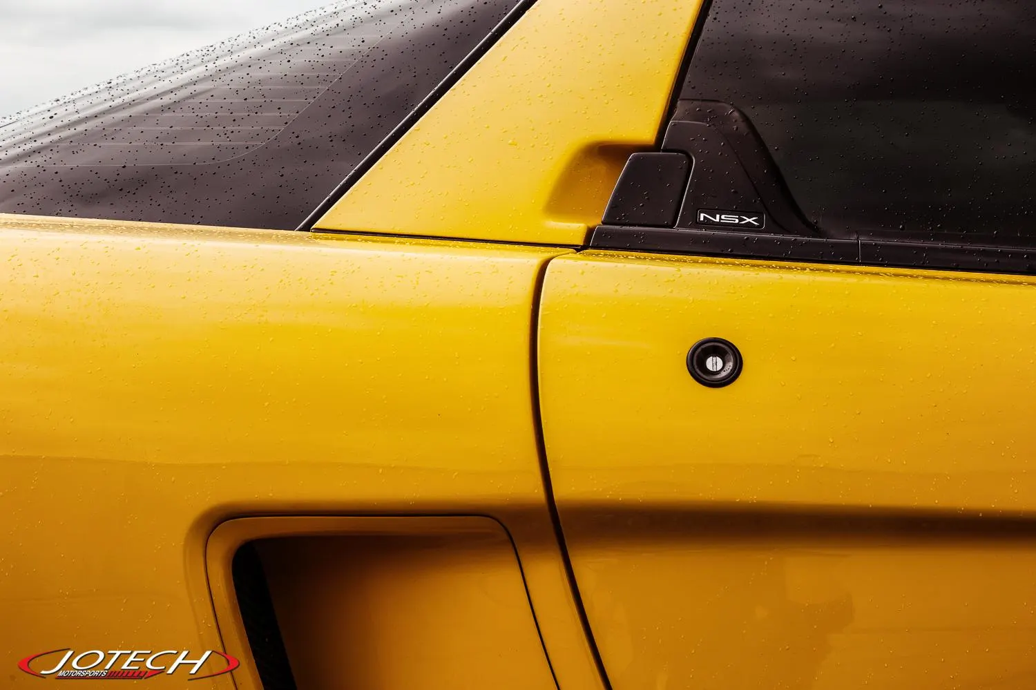 Close-up detail of a wet yellow Acura NSX sports car, showing the side panel, door, and rear window with water droplets.
