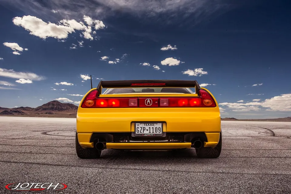 Rear view of a yellow Acura NSX with an aftermarket exhaust and large rear wing on a salt flat under a blue sky.