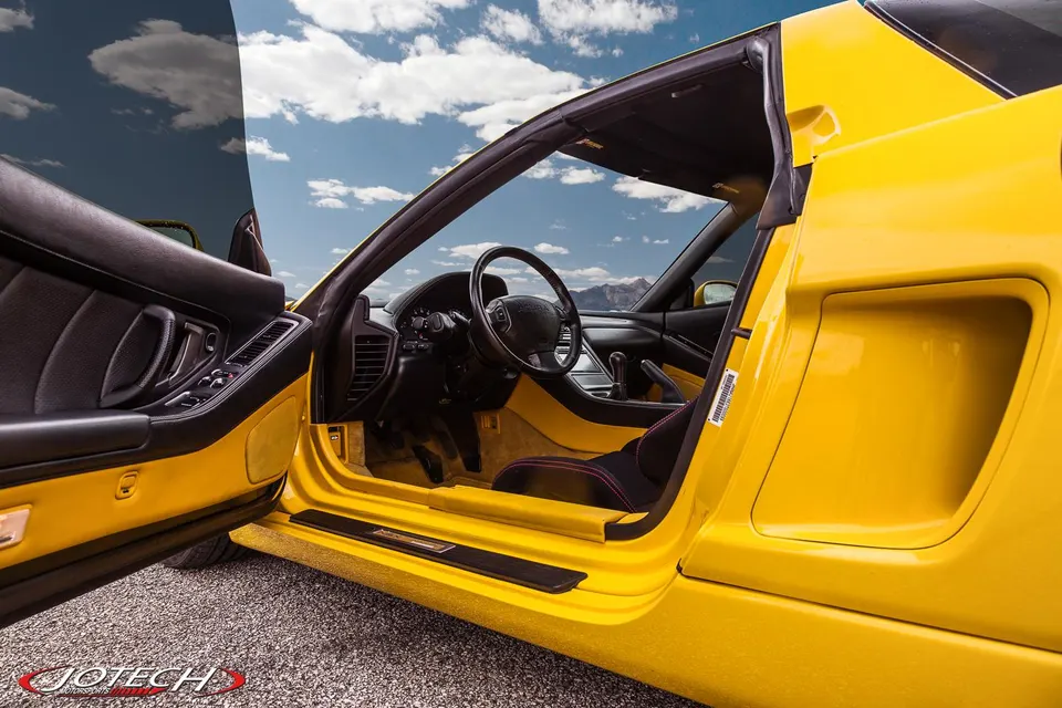 A yellow Acura NSX with the driver's door open, revealing the black interior, dashboard, and steering wheel under a blue sky.