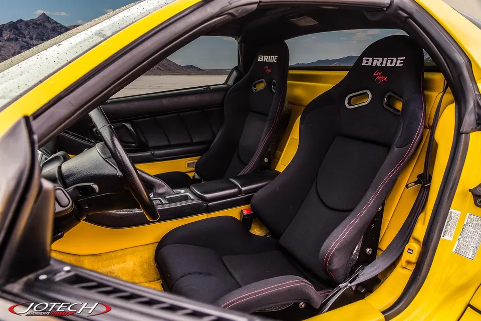 Interior of a yellow sports car featuring black Bride Low Max racing seats with red stitching, set against a desert landscape.