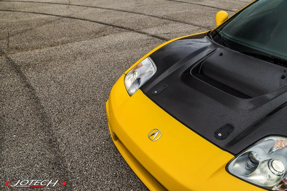 Yellow Acura NSX with a carbon fiber hood featuring a prominent scoop and hood pins, parked on an asphalt surface.