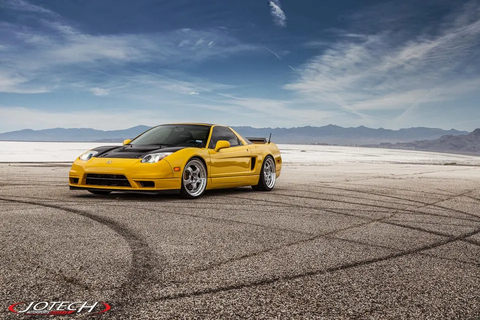 A vibrant yellow Honda NSX with a black hood and aftermarket wheels parked on a dry lake bed with mountains in the background.