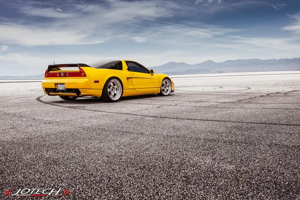 Yellow Acura NSX with aftermarket wheels and a large rear wing parked on an open road with tire marks.