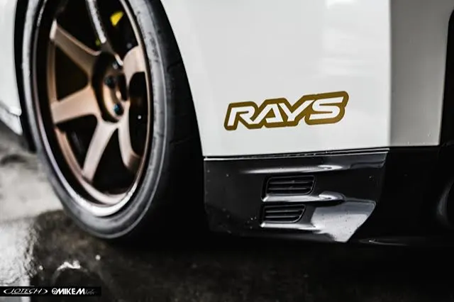 Close-up of a white car's rear fender featuring bronze RAYS wheels and a performance tire, highlighting the wheel and body detail.