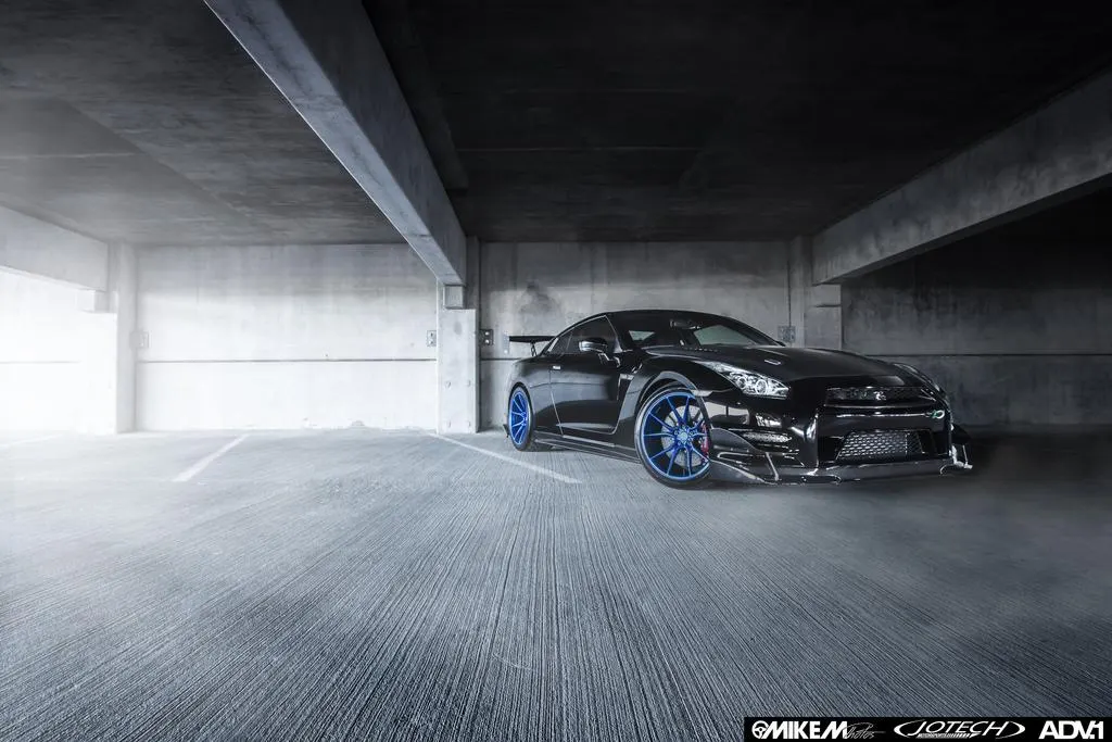 Black Nissan GT-R with blue aftermarket wheels and aero kit parked dramatically in a concrete parking garage.