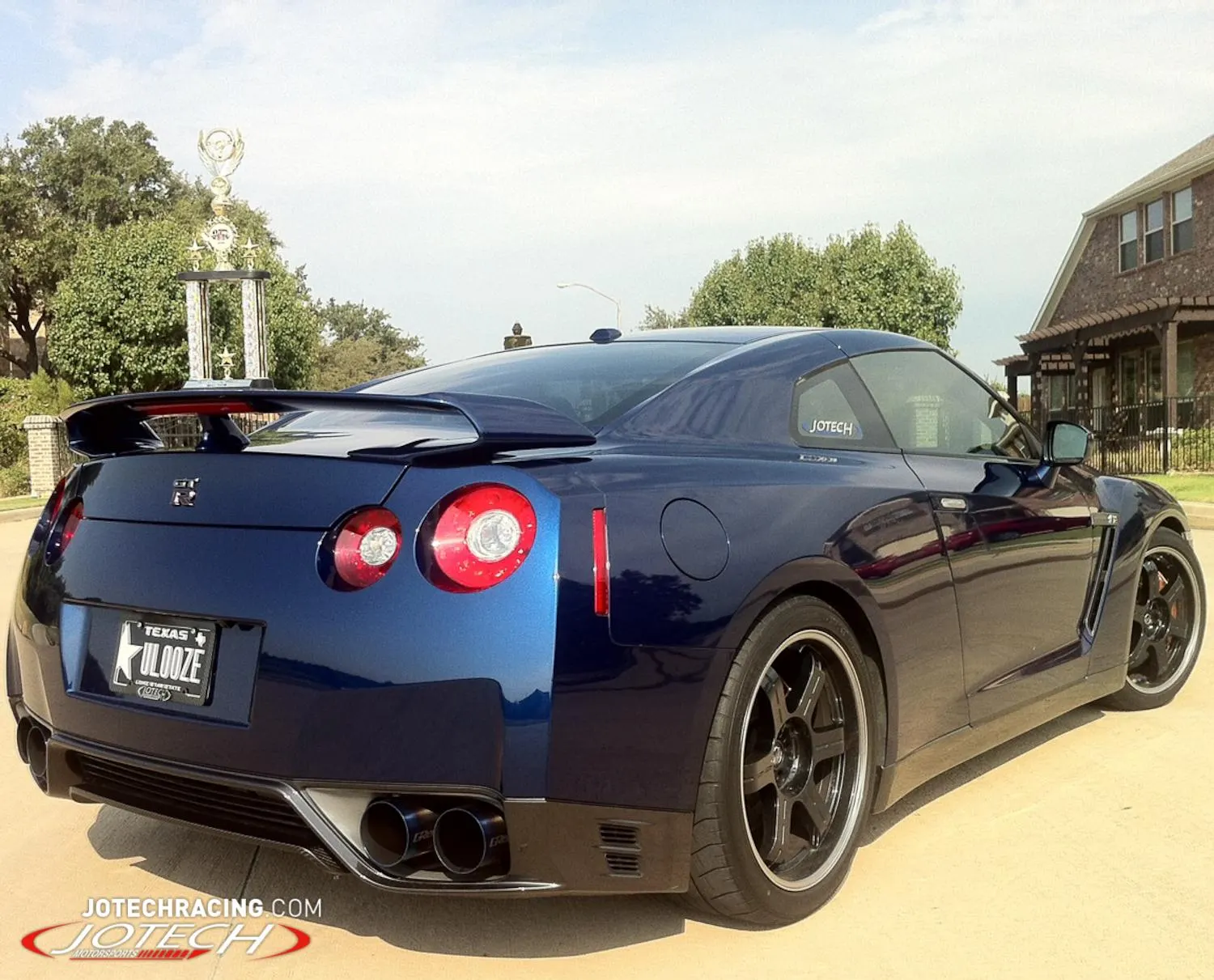 A blue Nissan GT-R with aftermarket wheels and exhaust, parked outdoors with a trophy in the background.