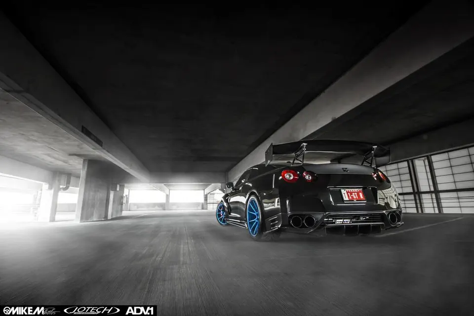 Rear view of a black Nissan GT-R with a large carbon fiber wing, aggressive diffuser, and blue aftermarket wheels in a parking garage.