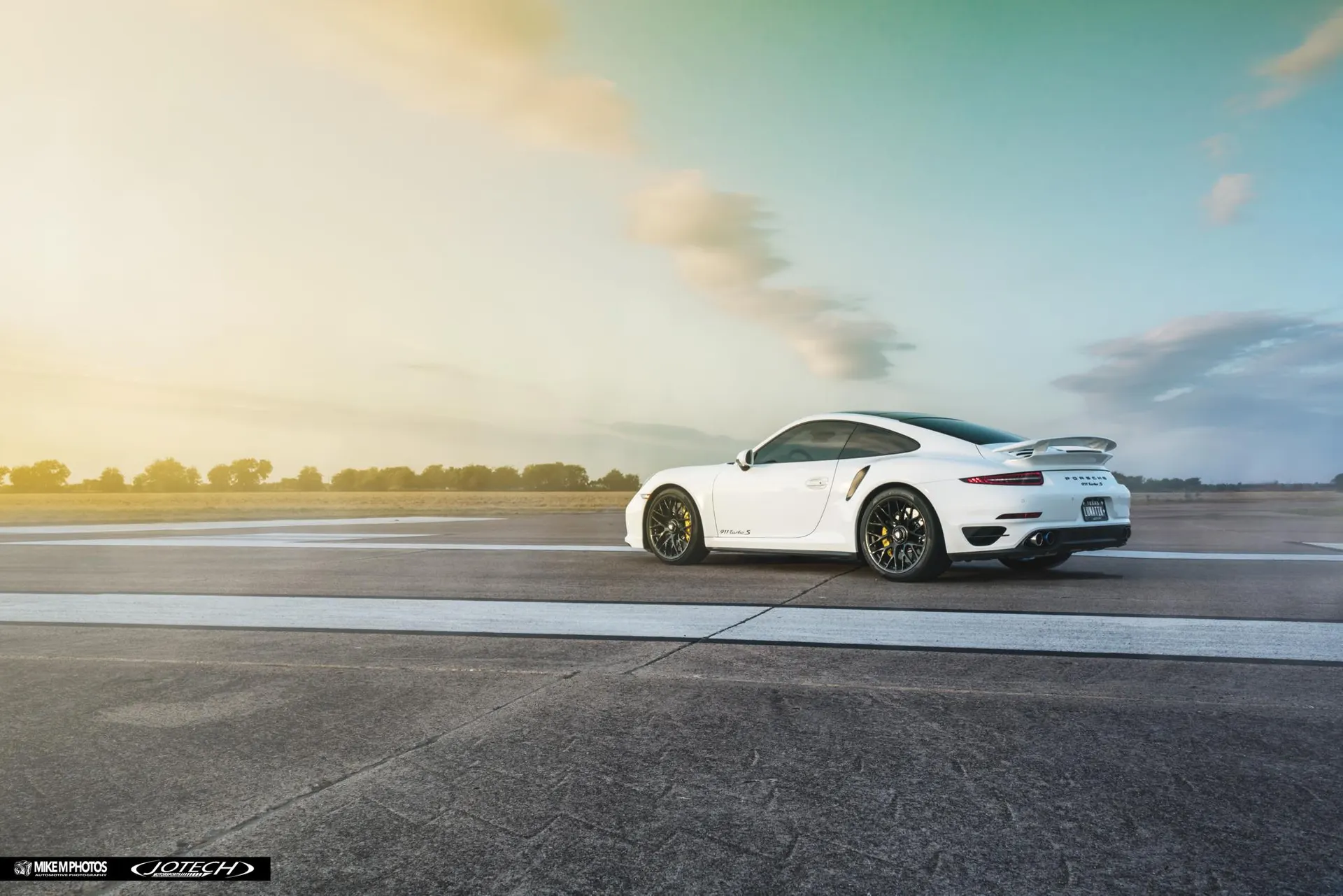 A white Porsche 911 Turbo S parked on an airfield runway at sunset, showcasing its sleek design and rear wing.