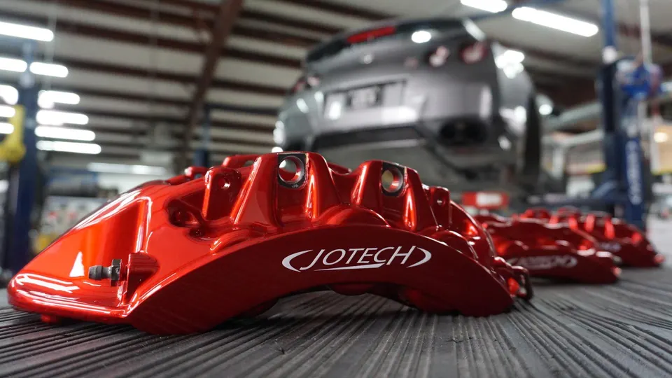 Red Jotech performance brake calipers on a workbench with a Nissan GT-R on a lift in the background at a tuning shop.