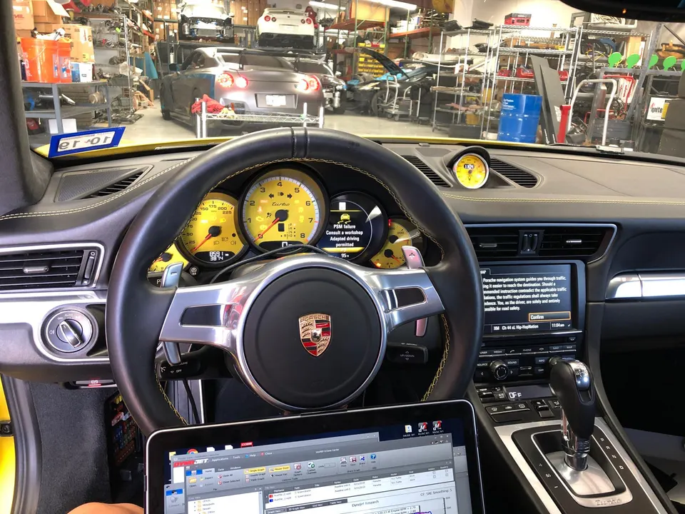 A Porsche 911 interior with a laptop connected for tuning, showing the dashboard and a performance shop in the background.