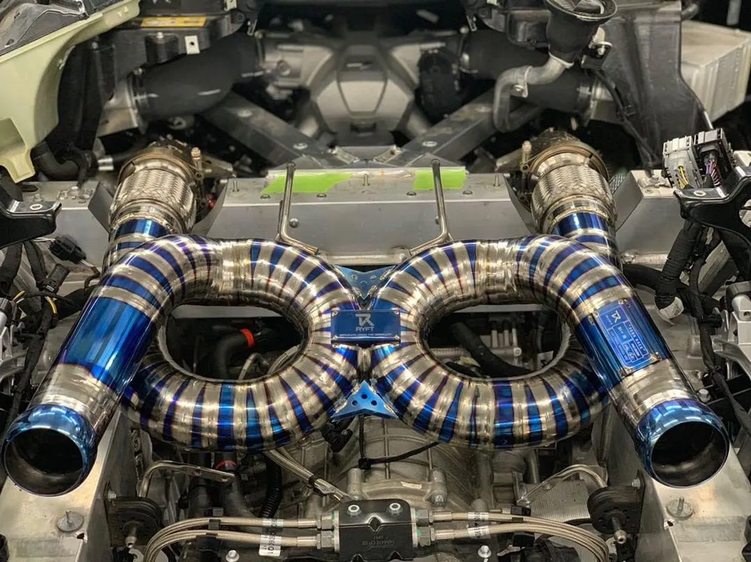 Close-up of a custom titanium turbo exhaust system in a high-performance car's engine bay, featuring blue and silver heat-treated piping.