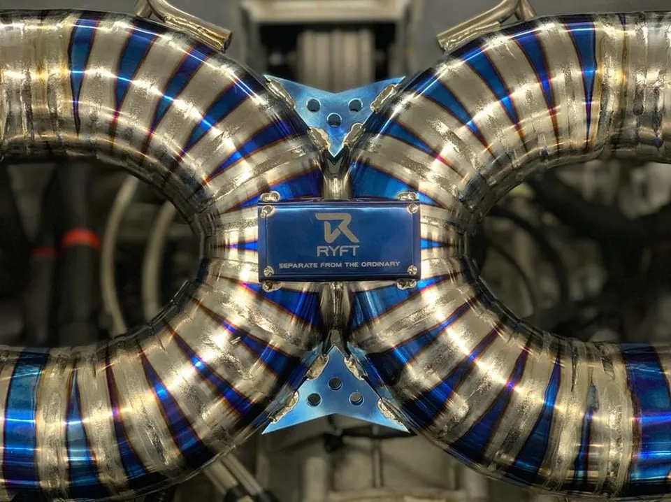 Close-up of a RYFT titanium exhaust system with intricate welds and blue heat-treated piping, showcasing performance car parts.