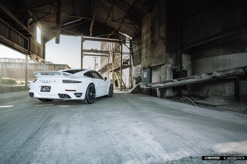 A white Porsche 911 Turbo S with aftermarket wheels and a rear wing parked in an abandoned industrial building.