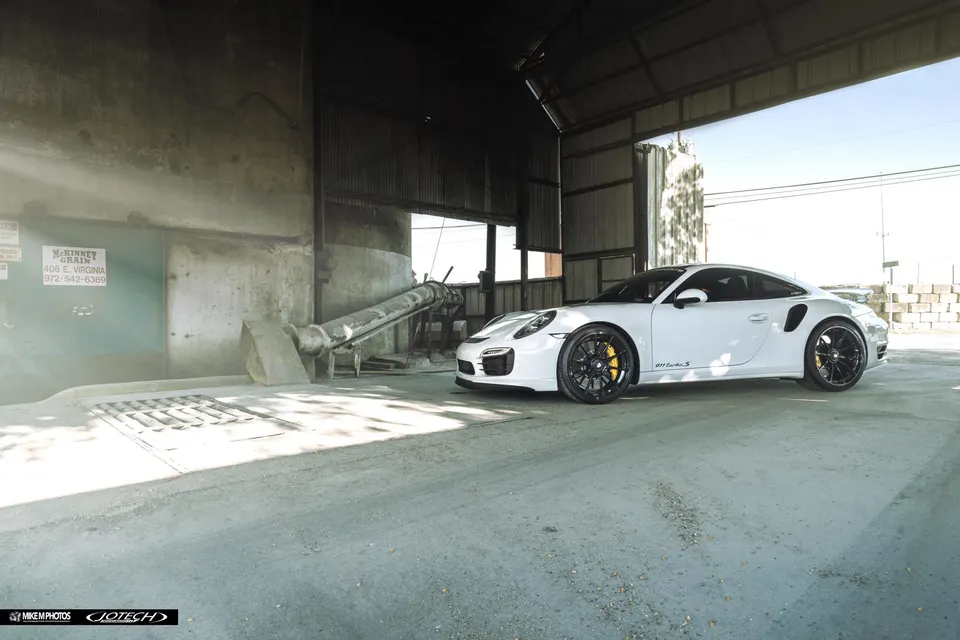 A white Porsche 911 Turbo S with black aftermarket wheels and yellow brake calipers parked in an industrial building.