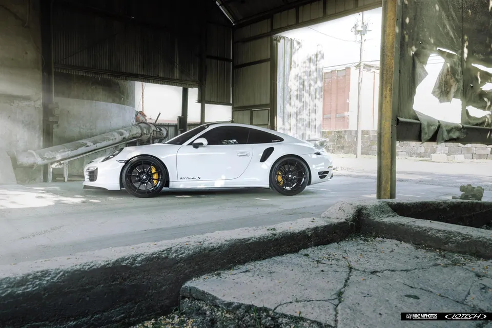 A white Porsche 911 Turbo S with black wheels and yellow brake calipers parked in an industrial setting.