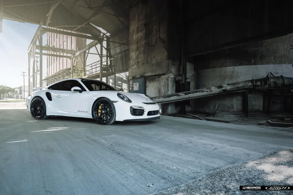 A white Porsche 911 Turbo S with black wheels and yellow brake calipers parked in an industrial setting.