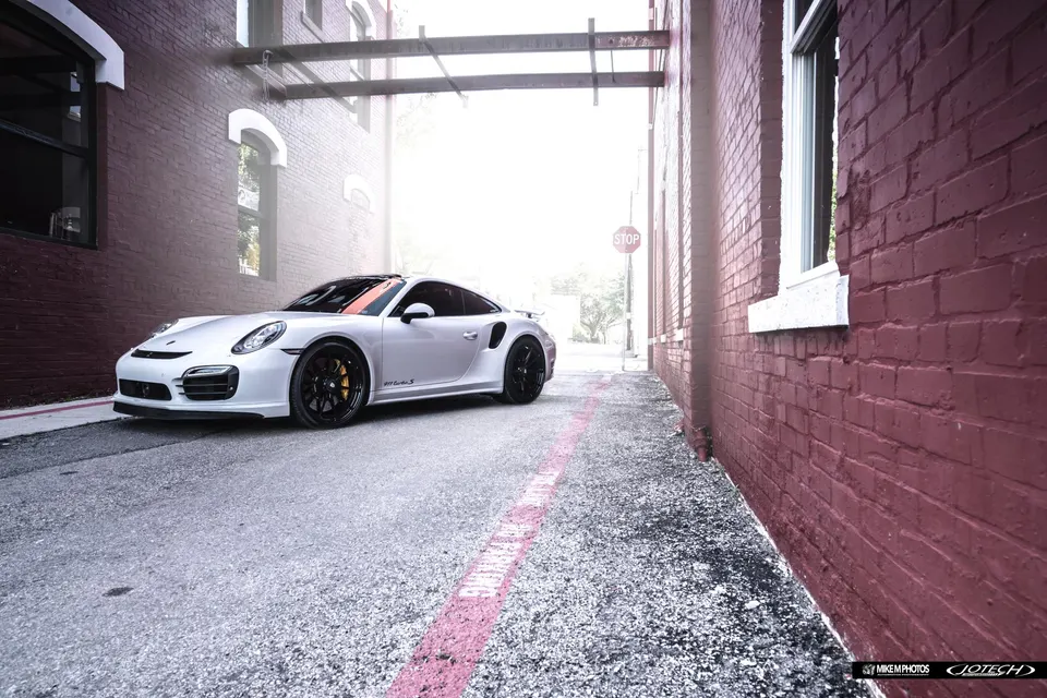 White Porsche 911 Turbo S with black aftermarket wheels parked in a brick alleyway during a photoshoot.