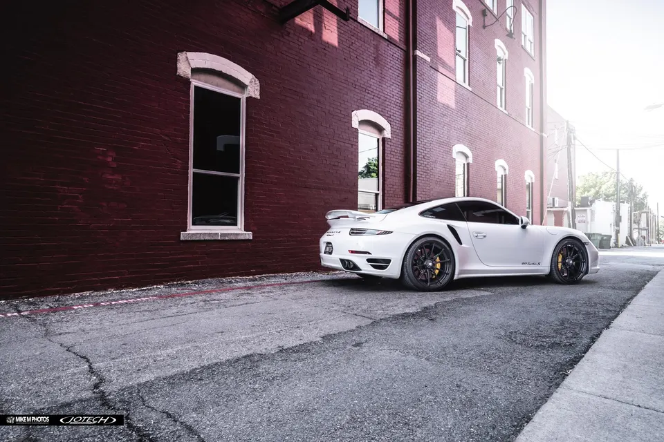 White Porsche 911 Turbo S with aftermarket wheels and a rear wing parked on a street next to a red brick building.