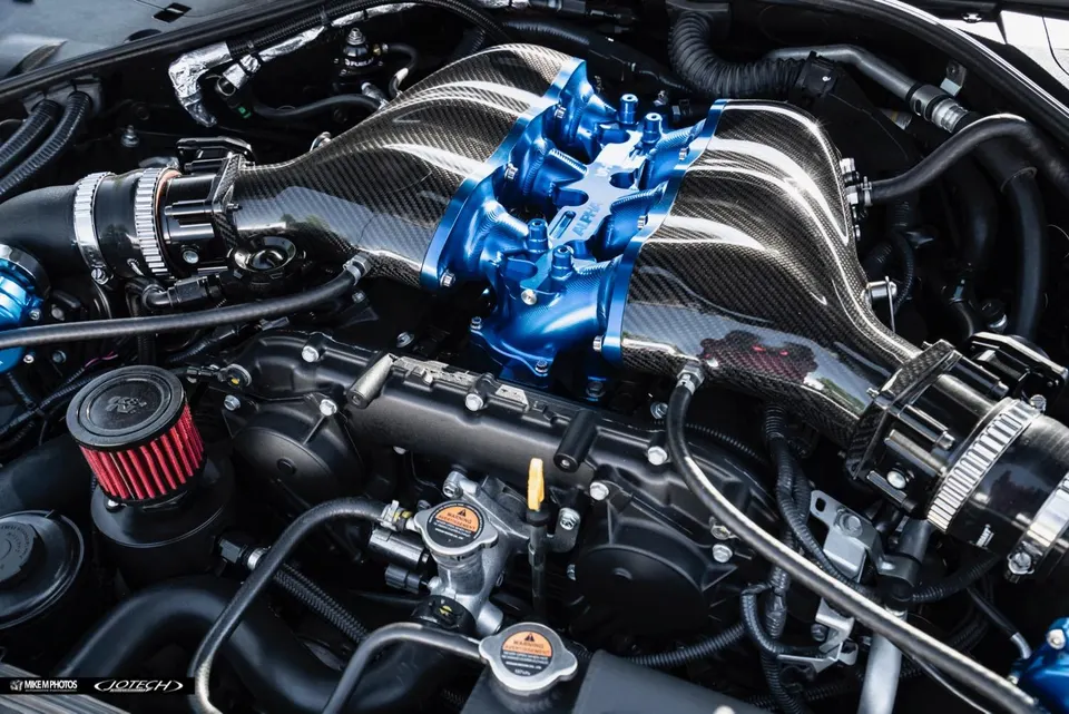 A close-up view of a custom engine bay featuring a carbon fiber intake manifold with a blue billet plenum and an aftermarket air filter.