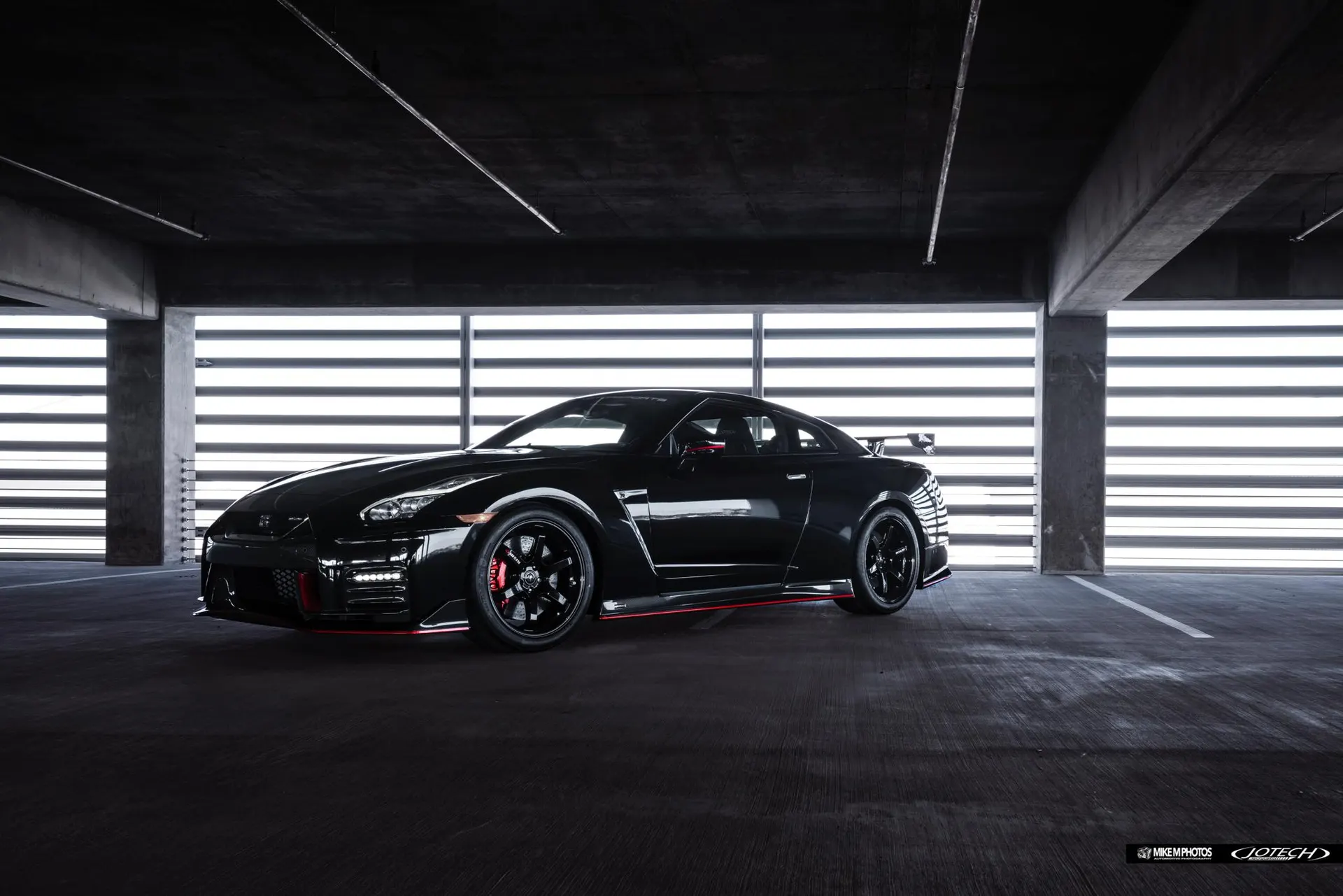 Black Nissan GT-R with aftermarket wheels and aero kit parked in a modern garage with striped lighting.