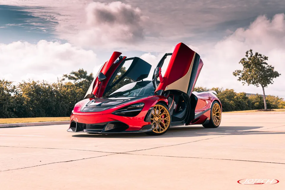 A red McLaren 720S with butterfly doors open, featuring gold aftermarket wheels, parked outdoors under a cloudy sky.