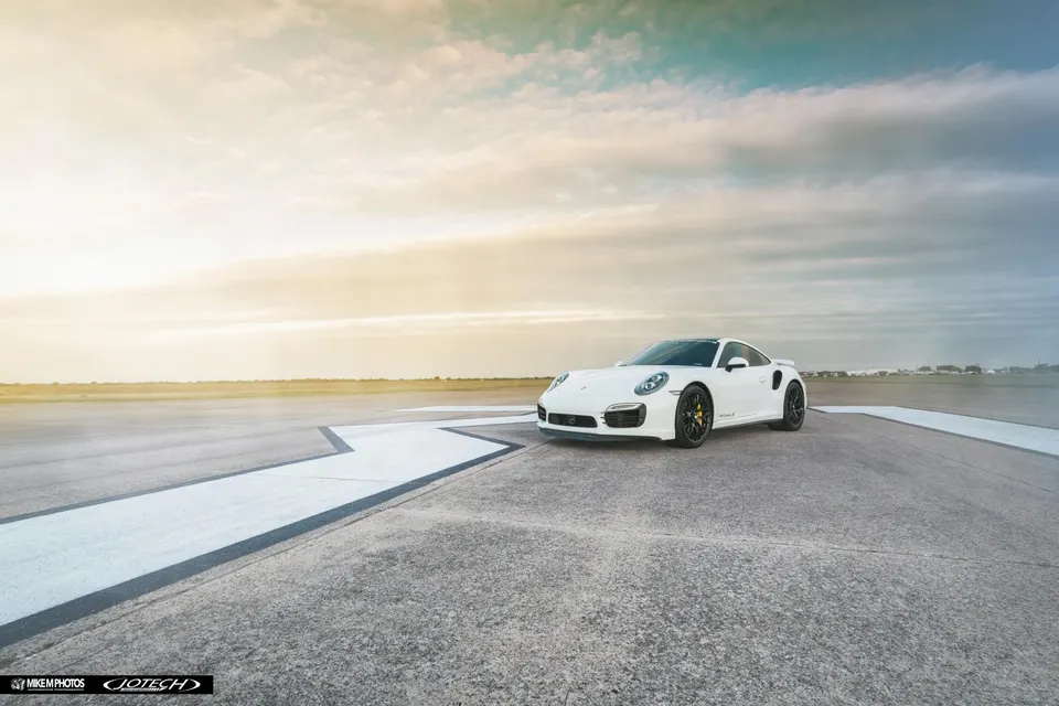 White Porsche 911 Turbo S parked on an airfield runway at sunset, showcasing its sleek design.