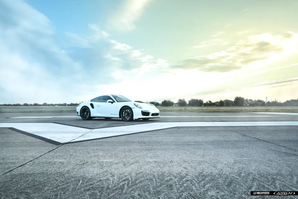 White Porsche 911 Turbo S sports car parked on an airfield at sunset, showcasing its sleek design and aftermarket wheels.