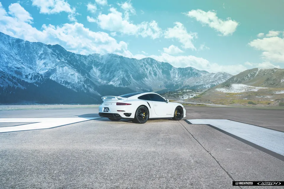 A white Porsche 911 Turbo S parked on an asphalt road with a majestic mountain range under a blue sky.