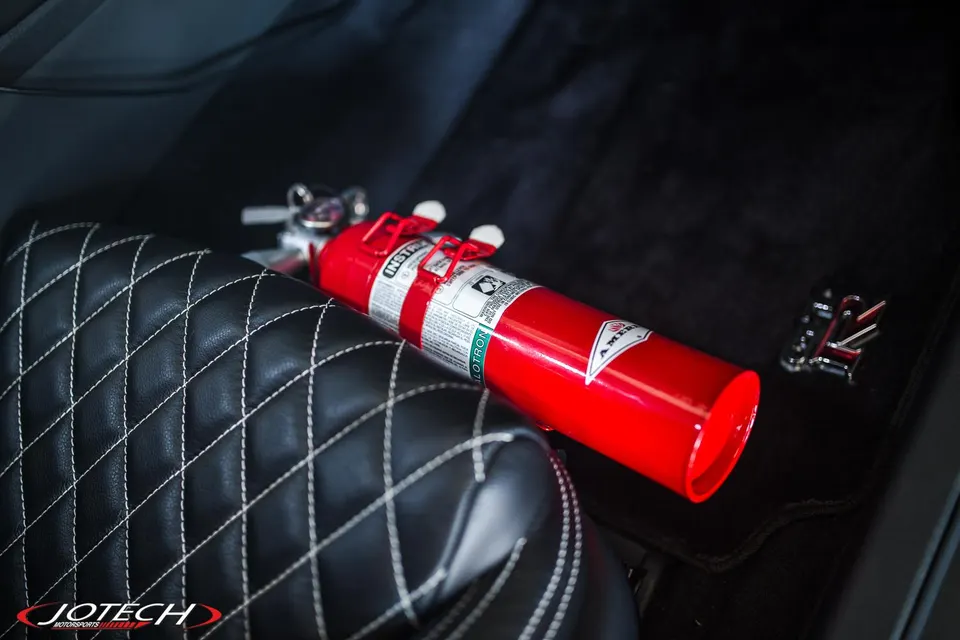 Red fire extinguisher mounted in a Nissan GT-R interior with quilted leather seats and GT-R floor mat.