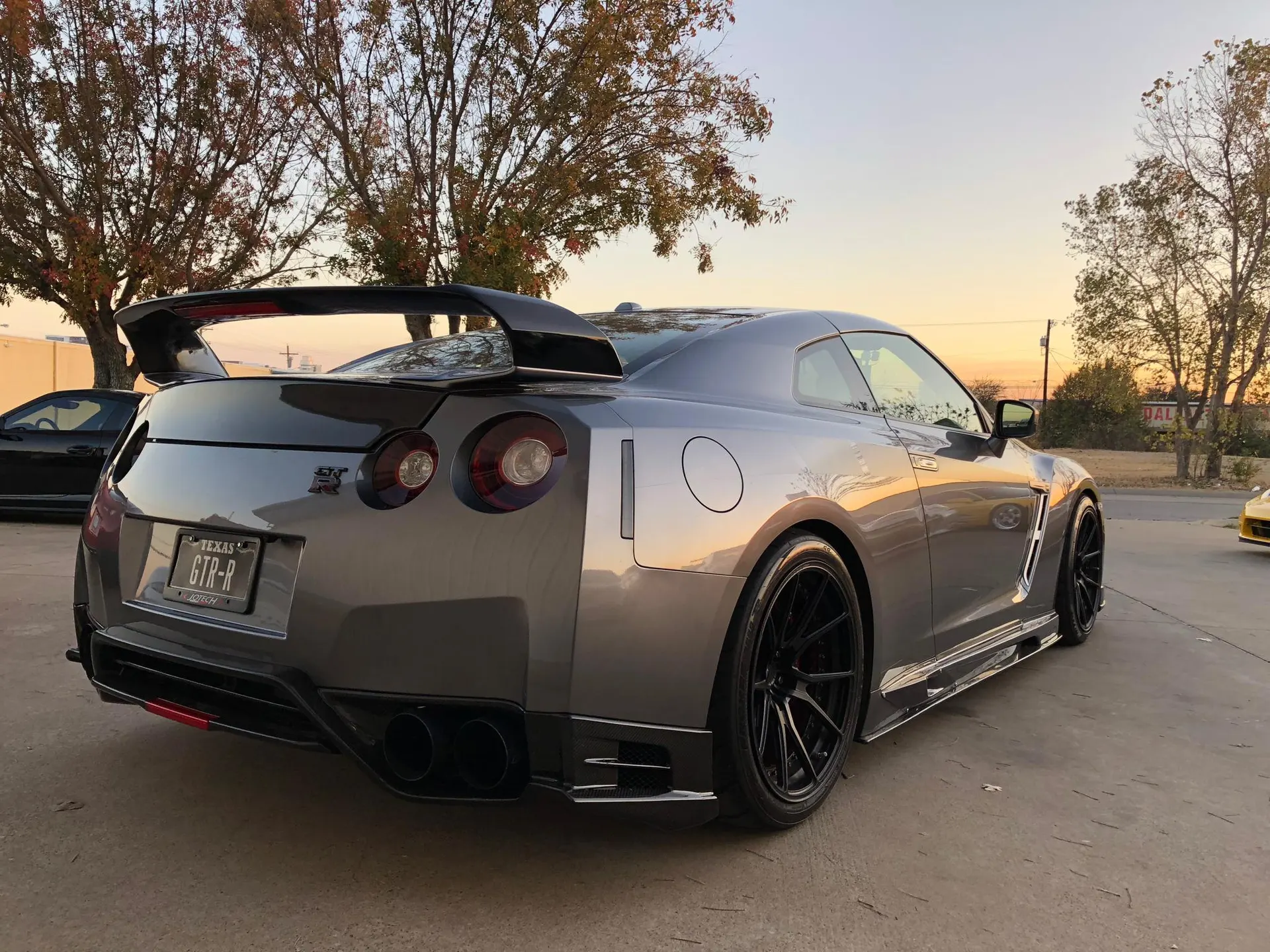 A grey Nissan GT-R with aftermarket wheels and a large rear wing parked outdoors at sunset.