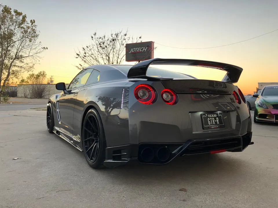 Rear view of a grey Nissan GT-R with a large carbon fiber wing, aftermarket wheels, and custom exhaust tips at sunset.
