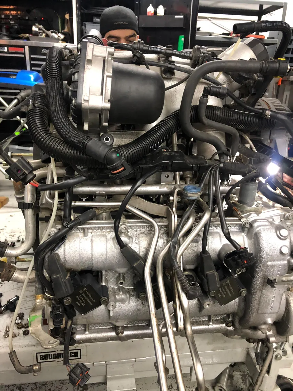 Mechanic working on a car engine assembly, showing the intake manifold, fuel lines, and ignition coils in a shop.