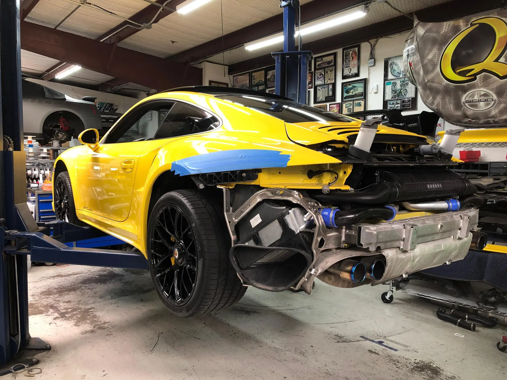 A yellow Porsche 911 on a lift in a shop with its rear bumper removed, revealing the exhaust and intercooler system.