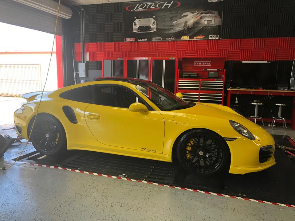 A bright yellow Porsche 911 Turbo strapped to a dynamometer at a performance tuning shop, ready for power testing.