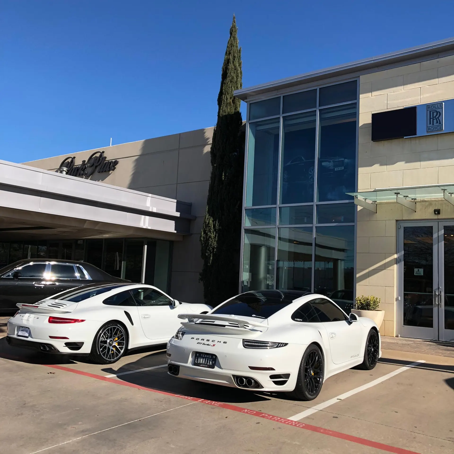 Two white Porsche 911 Turbo S sports cars parked outside a luxury car dealership on a sunny day.
