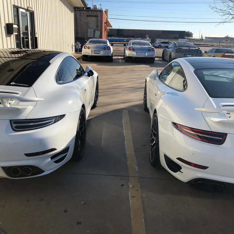 White Porsche 911s and silver Nissan GT-Rs parked outside a performance tuning shop on a sunny day.