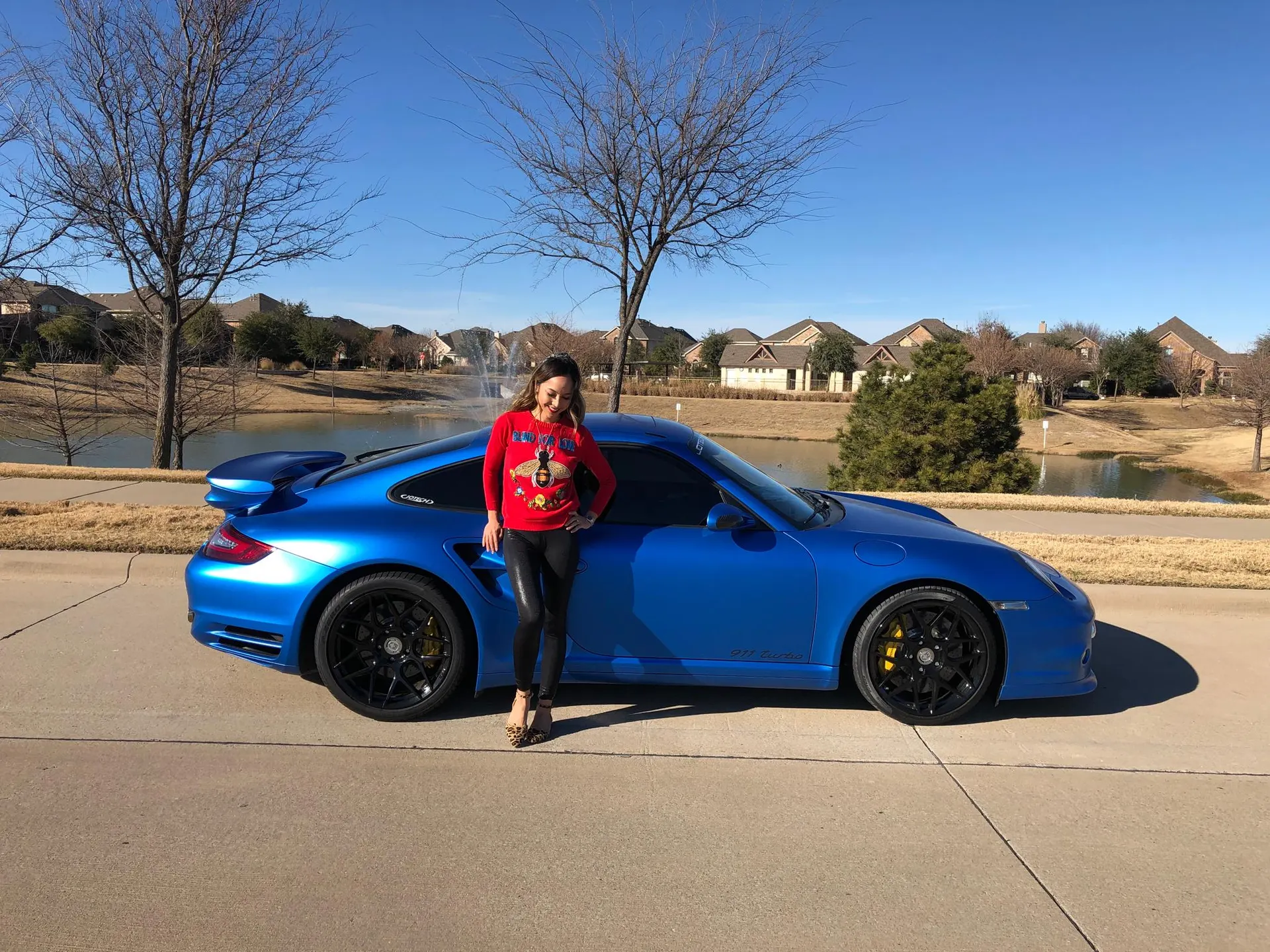 A woman in a red sweater stands next to a custom blue Porsche 911 Turbo with black wheels in a residential area.