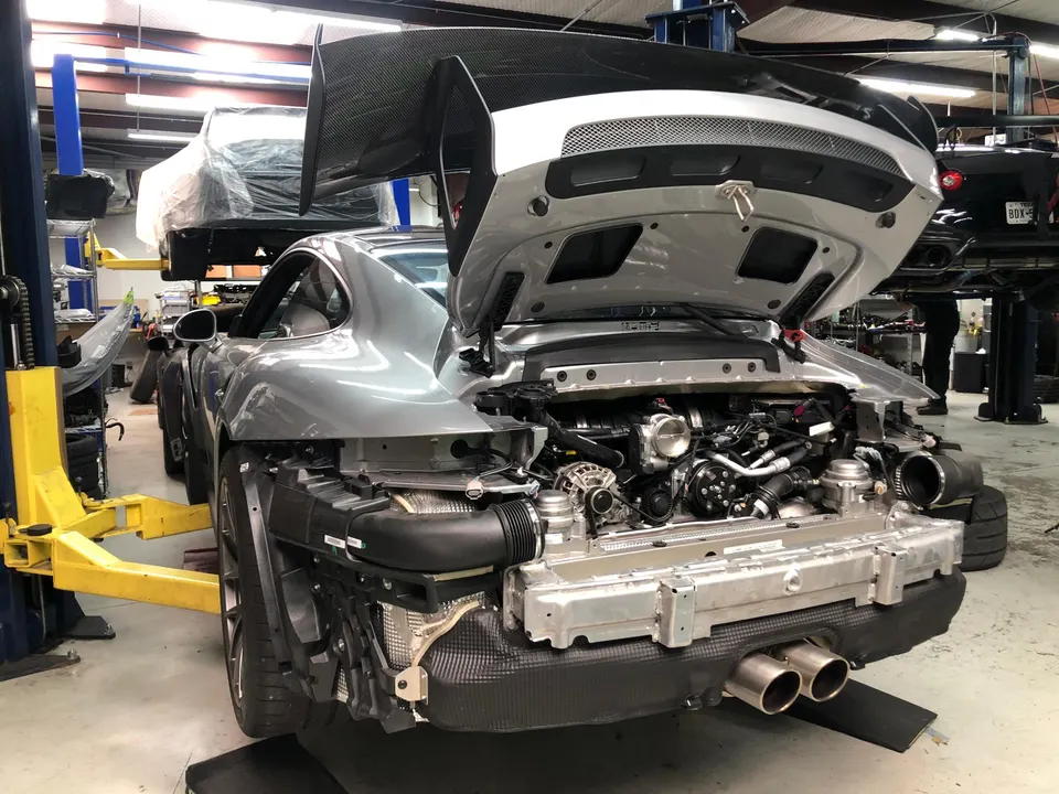 A grey Porsche 911 with its rear bumper removed, exposing the engine and a large carbon fiber wing, in a performance shop.