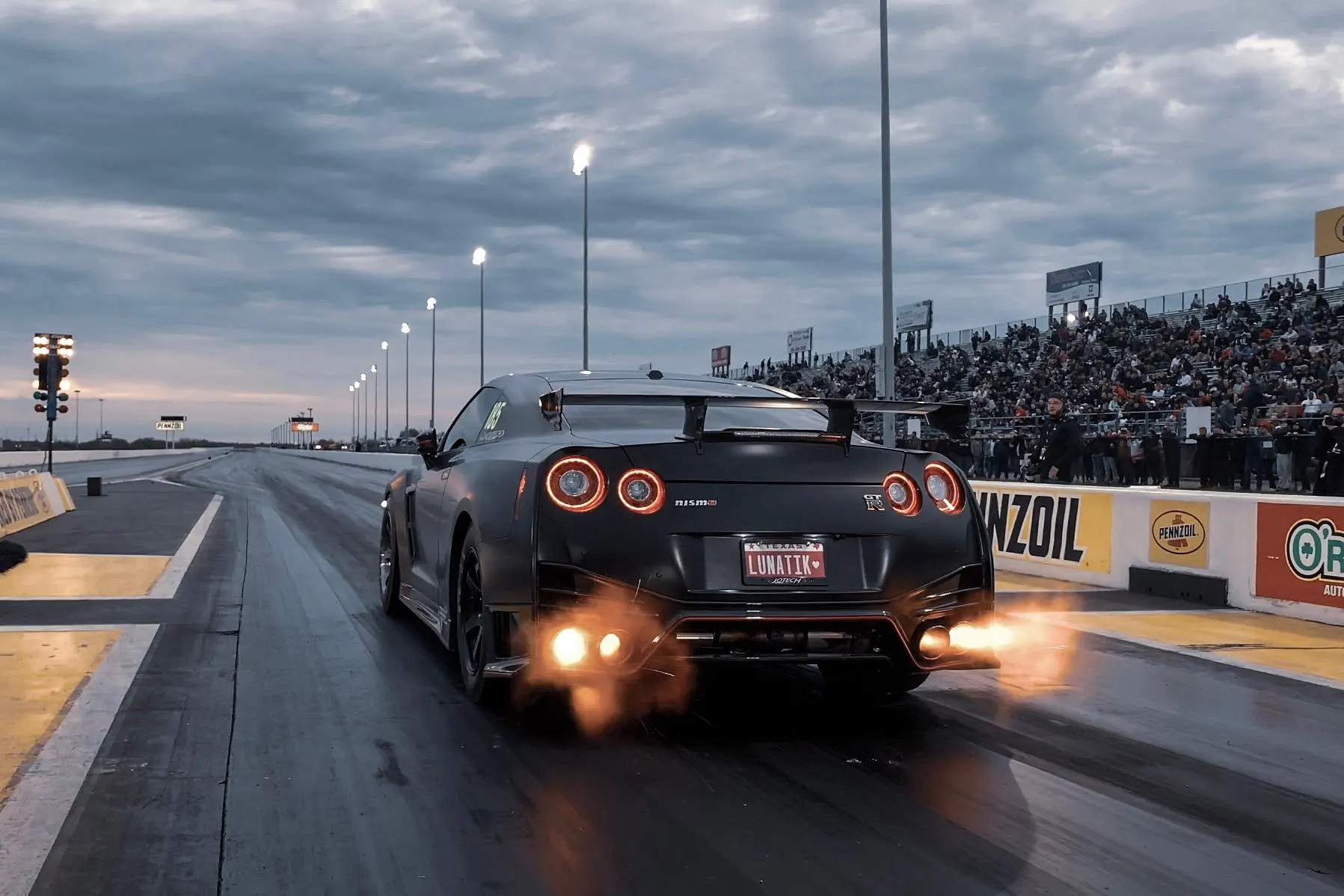 Black Nissan GT-R with exhaust flames launching on a drag strip with spectators in the grandstands.
