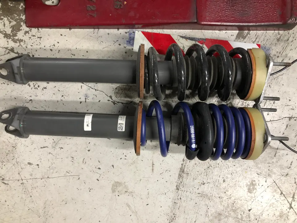 Two suspension struts with springs, one with blue aftermarket springs, on a shop floor.