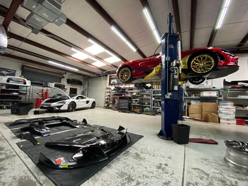 Red McLaren 720S on a lift and a white McLaren with its hood open in a performance tuning shop, with carbon fiber aero parts on the floor.