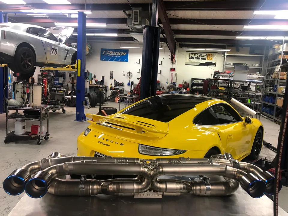 A yellow Porsche 911 Turbo and a silver Nissan GT-R on lifts in a performance tuning shop, with an aftermarket exhaust system in the foreground.