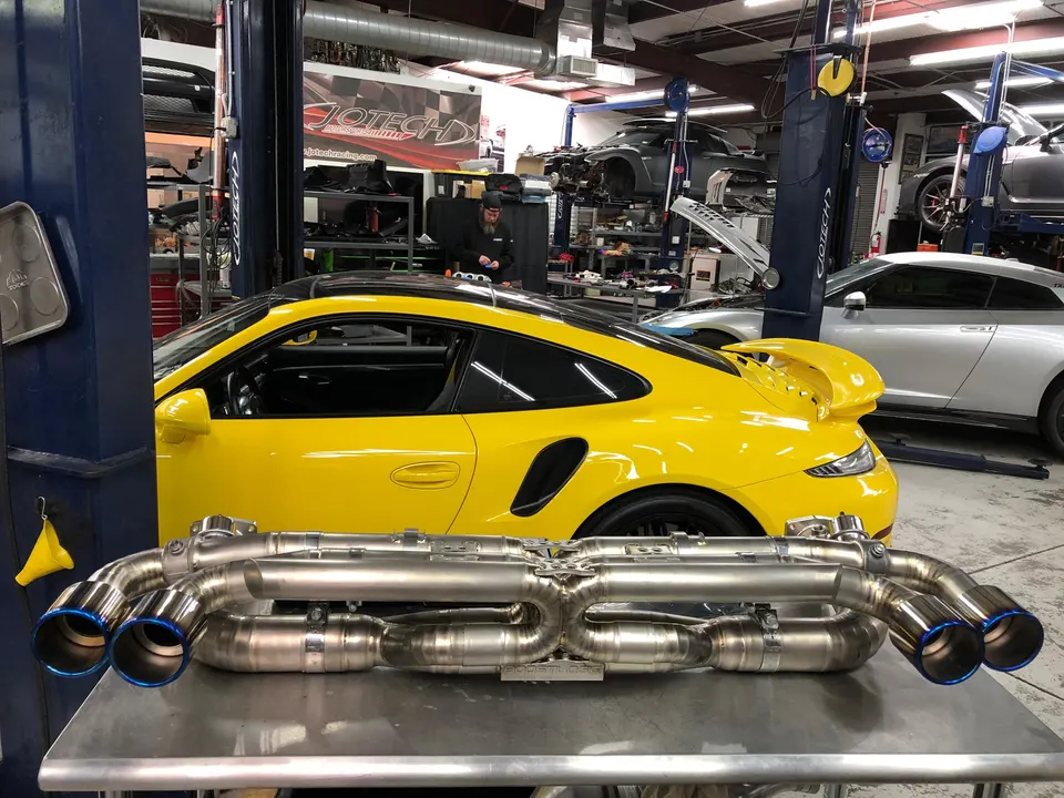 A yellow Porsche 911 in a Jotech Motorsports shop with a new titanium exhaust system on a table in the foreground.