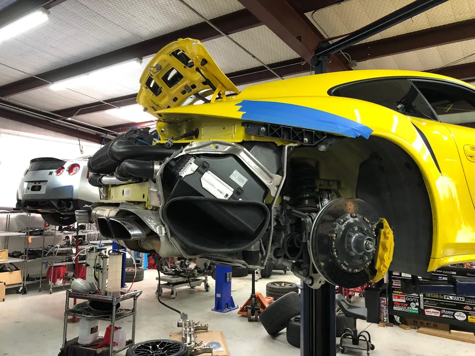 Yellow Porsche 911 GT3 RS on a lift in a performance shop with rear bumper removed and front wheel off, showing brakes.