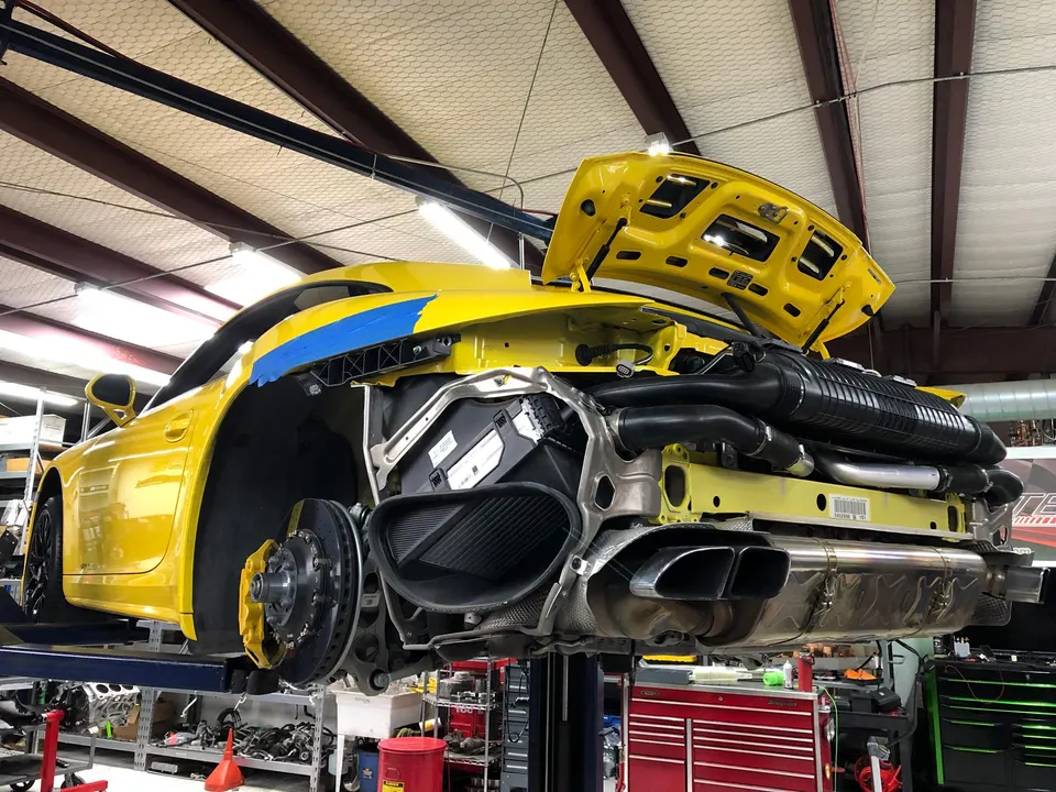 A yellow Porsche 911 on a lift in a performance shop, with its rear bumper removed, exposing the exhaust system and intercooler.