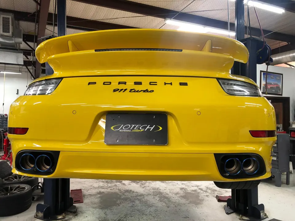 A yellow Porsche 911 Turbo on a lift in a Jotech Motorsports shop, showcasing aftermarket exhaust tips.