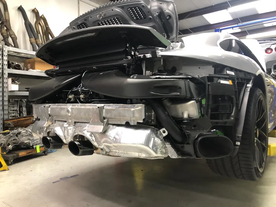 A Porsche 911 in a shop with its rear bumper removed, revealing the exhaust system and engine components.